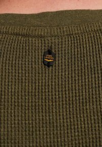 Tissu de pull à côtes vert avec une étiquette noire portant "PME LEGEND" en orange, fixée au col.