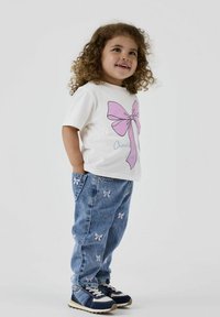 Weißes T-Shirt mit großem pinkfarbenem Schleifen-Design, kombiniert mit blauen Jeans aus Denim, die kleine gestickte Schleifen aufweisen. Schwarze und weiße Sneakers runden das Outfit ab.