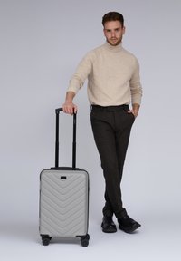 Jongeman in een beige coltrui en donkere broek staat met één hand in zijn zak en de andere hand die een zilveren koffer met wielen vasthoudt.
