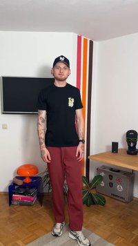 Črna grafična majica z rumenim motivom, rdeče hlače in sive superge. V okolici so televizor, oranžna luč in lesena miza.