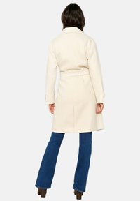 Beige, knee-length coat with a belted waist, notched collar, and buttoned cuffs; model wearing blue flared jeans and heeled boots.