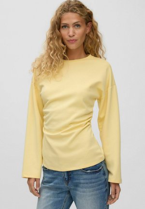 Kvinde med krøllet blondt hår iført en lysegul langærmet top og blå jeans, står mod en ensfarvet lys baggrund.