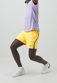 Kortärmad lila träningsskjorta med orange detaljer, i kombination med gula shorts med siddragkedja och svart Nike-logga, bärs med vita skor.