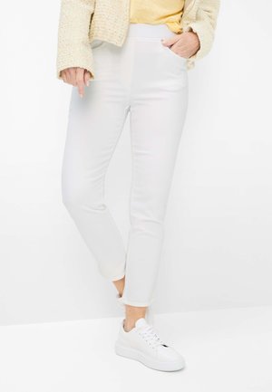 Person iført hvide tætsiddende bukser, hvide sneakers, en gul top og en beige tekstureret jakke, med den ene hånd i lommen.