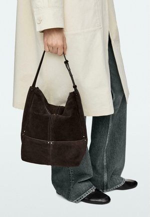 Persona che indossa un cappotto beige chiaro e jeans larghi scuri tiene una grande borsa in camoscio marrone scuro con manico corto.