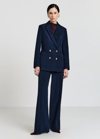 Blazer doppiopetto in velluto a coste blu navy con due grandi bottoni, abbinato a pantaloni a gamba larga coordinati. Completato da un dolcevita scuro.