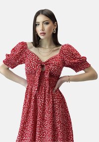 Rotes Blumenkleid mit kurzen Puffärmeln, gerafftem Oberteil und einer gebundenen Schleife vorne. Aus leichtem, gemustertem Stoff gefertigt.