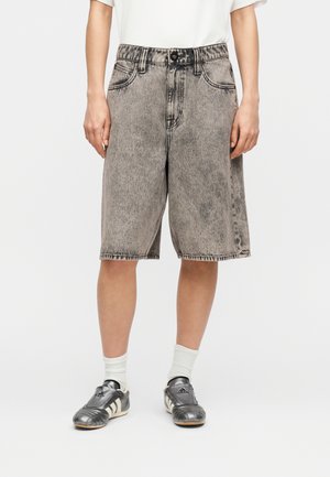 Persoană purtând pantaloni scurți lungi din denim gri spălat acid, șosete albe până la gleznă, adidași Adidas negri și albi și un tricou alb, stând pe un fundal alb.