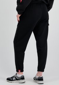 Schwarze Jogginghosen mit einem elastischen Bund, Bündchen an den Knöcheln und bedrucktem "ALPHA"-Logo an der Seite. Kombiniert mit schwarzen und grauen Sneakern.