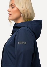 Marinblå huvjacka i mjukt tyg, med en profilbild, justerbar dragsko och ett svart logotyp-märke på ärmen.