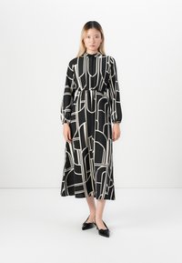 Jeune femme portant une robe midi à manches longues à motifs géométriques noirs et blancs avec des talons bas noirs, se tenant contre un mur blanc.