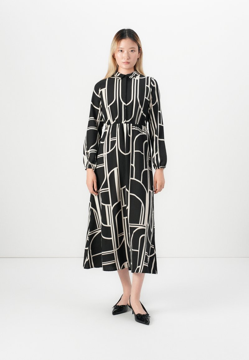 Jeune femme portant une robe midi à manches longues à motifs géométriques noirs et blancs avec des talons bas noirs, se tenant contre un mur blanc.