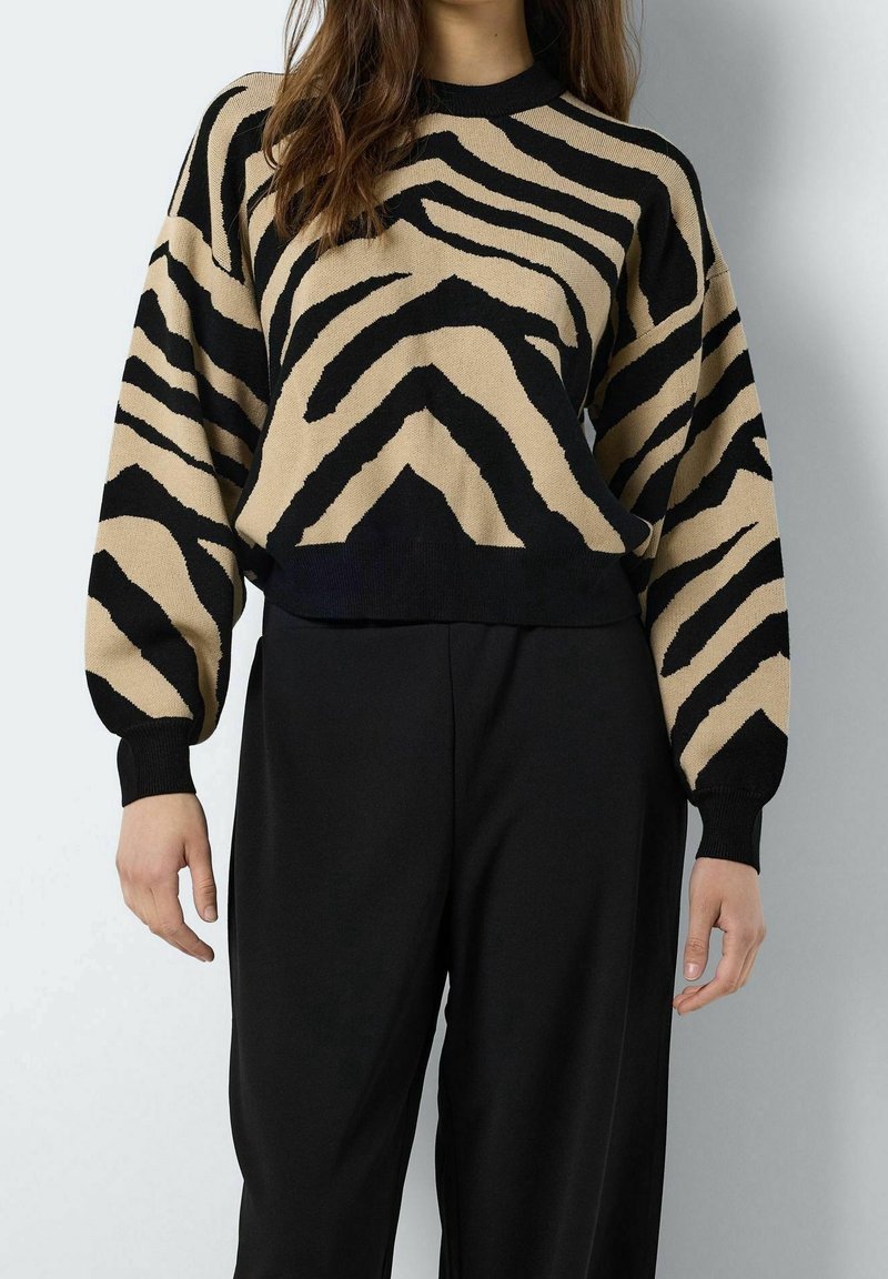 Femme portant un pull à rayures zèbre beige et noir avec un pantalon noir à jambes larges sur un fond clair uni.