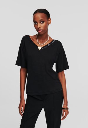 T-shirt nera con scollo a V e maniche corte, tessuto morbido, vestibilità rilassata. Presenta un accento bianco sulla spalla e una collana a strati in argento.