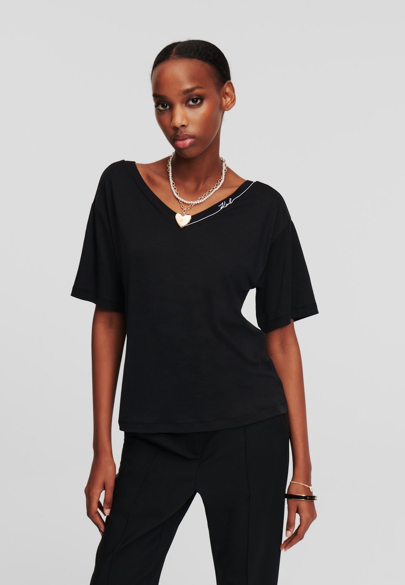 Tricou negru cu decolteu în V, mâneci scurte, dintr-un material moale, cu o croială lejeră. Prezintă un accent alb pe umăr și un colier din argint stratificat.