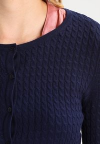 Cárdigan de punto azul marino con diseño de cable texturizado, cuello redondo y botones negros en la parte delantera. Se ve una camiseta rosa claro debajo.