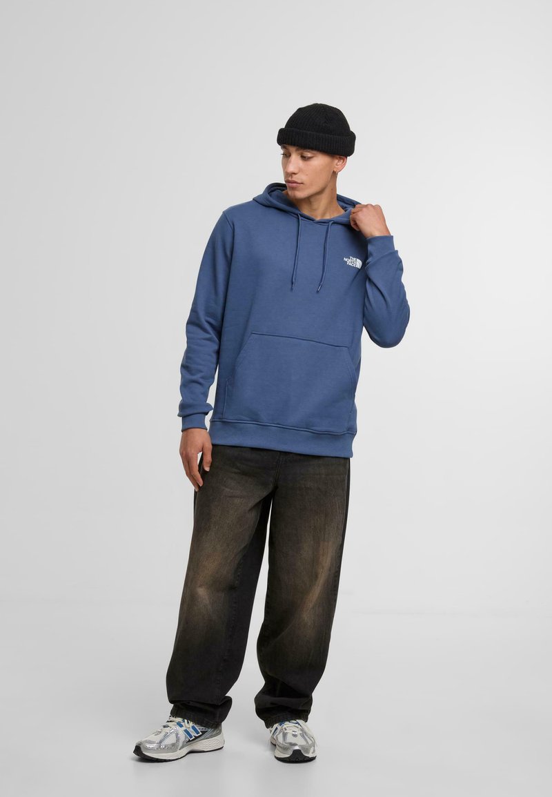 Blauwe hoodie met een voorzak en een trekkoordcapuchon, gecombineerd met losse, donkere jeans en zilveren hardloopschoenen. Zwarte gebreide muts gedragen.