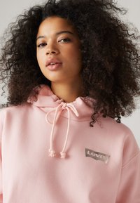Ljusrosa hoodie med dragsko på huvan, med en metallisk Levi's logotyp patch på bröstet. Mjuk tyg och avslappnad passform.