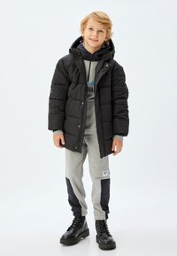 Svart vadderad jacka med huva, grå sweatshirt under, grå sweatsbyxor med blå och svarta accenter, och svarta kraftiga kängor.