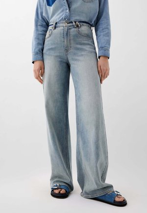 Jeans large bleu clair à taille haute, avec des coutures surpiquées et une fermeture à bouton à l'avant, associés à des sandales bleues à brides.