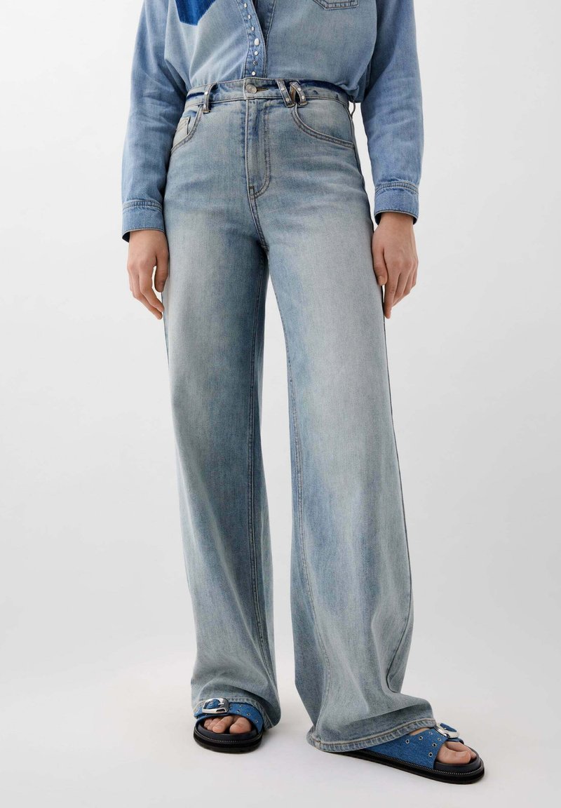 Jeans de culoare albastru deschis, cu croială largă și talie înaltă, având cusături decorative și închidere cu nasture în față, asortați cu sandale albastre cu bretele.