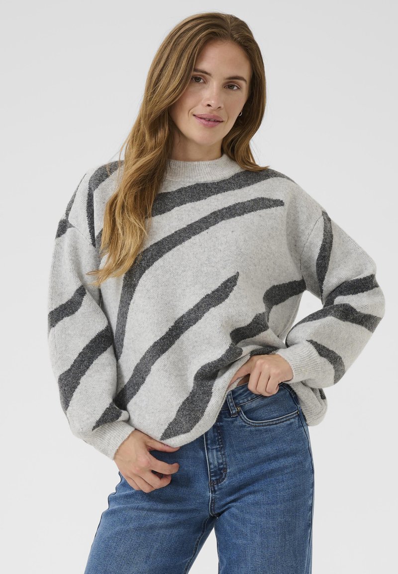 Jeune femme avec de longs cheveux châtain clair portant un pull gris avec des rayures abstraites sombres et un jean bleu, se tenant devant un fond blanc.