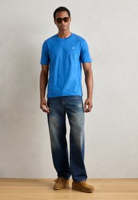 T-shirt de algodão azul com mangas curtas, gola redonda e pequeno logótipo. Combinado com jeans azuis desbotados e sapatos slip-on de camurça bege.