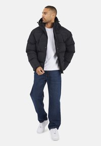 Doudoune noir avec un col haut, fermeture éclair et boutons-pression. Portée sur un t-shirt blanc et associée à un jean bleu et des baskets blanches.