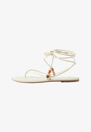 Flache weiße Sandale mit dünnen Riemen um den Knöchel und drei dekorativen Holzperlen nahe der Zehenschlaufe, beige Sohle mit abgestepptem Rand.