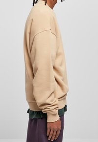 Beiges Sweatshirt mit Rundhalsausschnitt, lockerem Schnitt, gerippten Bündchen und weicher Textur, darüber ein dunkelgrünes Hemd und lilafarbene Hose getragen.