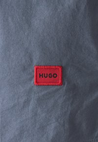 Grått tyg med en slät textur, med en röd broderad etikett med "HUGO" i svarta bokstäver. Designen är enkel och modern.