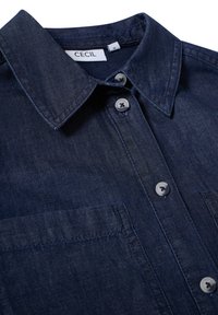 Mörk denimskjorta med krage, som har två framfickor, knappstängning och ljusgrå knappar. Slät textur, klassisk design.