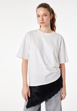 Ung kvinde med lysebrunt hår i en hestehale iført creoler, oversized hvid T-shirt, sort asymmetrisk nederdel over grå bukser, neutral udtryk.