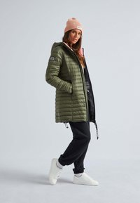 Olijfgroen pufferjack met capuchon, ritssluiting aan de voorkant en zijzakken; gecombineerd met zwarte broek en witte sneakers; lichtroze beanie op het hoofd.