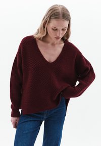 Bordeaux gebreide trui met een diepe V-hals, gestreept ribpatroon, relaxte pasvorm en lange mouwen. Gepaard met blauwe denim jeans.