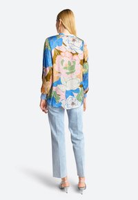Bunt bedruckte Bluse mit langen Ärmeln, die eine Mischung aus Blau, Pink, Grün und Brauntönen aufweist, kombiniert mit hellblauen Jeanshosen.