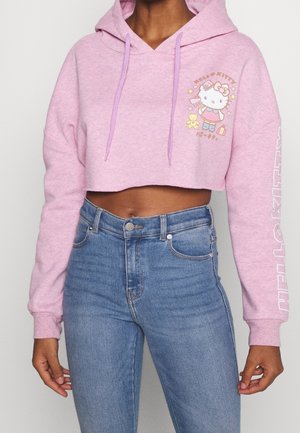 Femme portant un sweat à capuche court rose Hello Kitty avec un graphique à l'avant et du texte sur la manche, associé à un jean bleu taille haute.