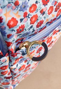 Prim-plan cu o geantă cu model floral cu flori roșii, mov și portocalii și un fermoar rotund auriu și negru.