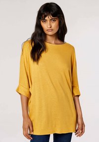 Tunique en tricot jaune moutarde avec une coupe décontractée, un encolure ronde et des manches trois-quarts, présentant une texture douce et un minimum de détails.