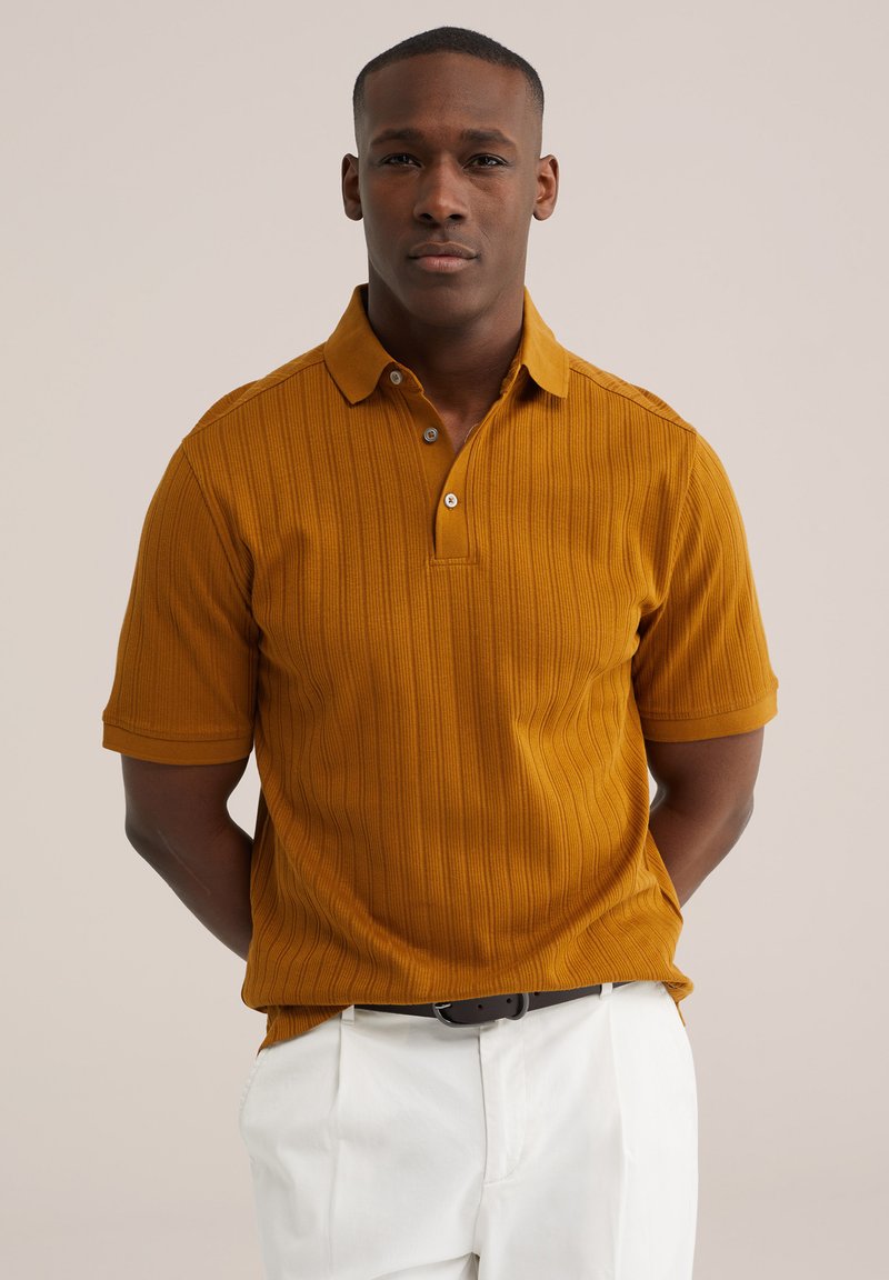 Uomo che indossa una polo a maniche corte a coste color senape con pantaloni bianchi e cintura nera, in piedi con le mani dietro la schiena.