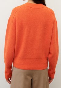 Orange stickad tröja med ribbat mönster, vida ärmar och en figurnära nederkant, i kombination med beige byxor. Enkel och varm design.