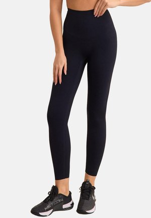 Kobieta w czarnych legginsach z wysokim stanem i czarnych butach sportowych, stojąca z jedną ręką opartą na biodrze.