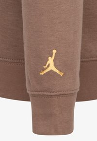 Rudos spalvos džemperio audinys su giliumis rankogaliais, kairiajame rankogalyje puikuojasi auksinė išsiuvinėta Jumpman logotipas. Lygi tekstūra, kasdieniškas dizainas.