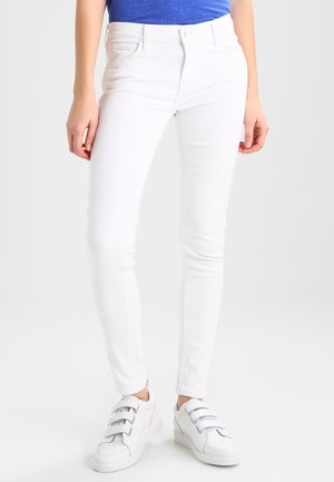 Femme portant un jean blanc slim, des baskets blanches à scratchs et un haut bleu tacheté sur un fond uni.