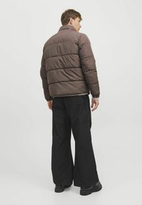 Giubbotto imbottito marrone con cuciture verticali, colletto alto e polsini elastici. Indossato sopra un maglione leggero e abbinato a pantaloni larghi neri.