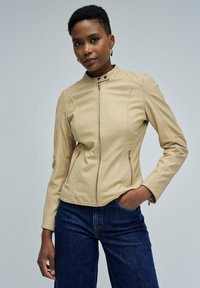 Femme aux cheveux courts et bouclés portant des boucles d'oreilles en or, une veste en cuir beige zippée et un jean bleu foncé, se tenant avec une main dans la poche.