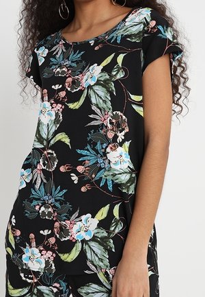 Femme portant un haut noir à manches courtes avec un motif floral bleu, rose et vert, assorti à un pantalon, sur un fond uni.