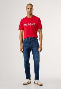 Rdeča majica s kratkimi rokavi s potiho "Pepe Jeans", v kombinaciji z modrimi kavbojkami in belimi športnimi copati s črnimi detajli.