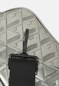 Borsa argentea con un motivo geometrico in rilievo, una tracolla nera con fibbia e il logo Lacoste ben in evidenza.