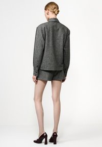 Szürke gyapjú keverék, oversize ing, gombos mandzsettával, hozzá illő sorttal párosítva. A modellen bordó magassarkú cipők vannak. Semleges háttér.